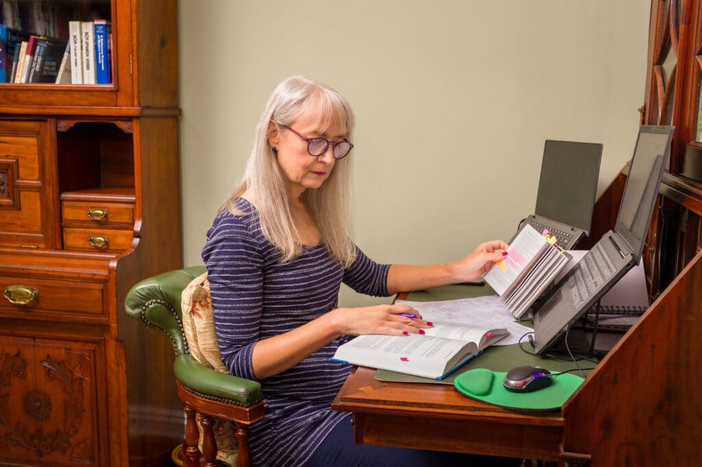 Melanie Cotton, proofreader and editor, gimlet-eyed grammar tickler® sitting at her desk, consulting hard copy resources while proofreading a document on her computer